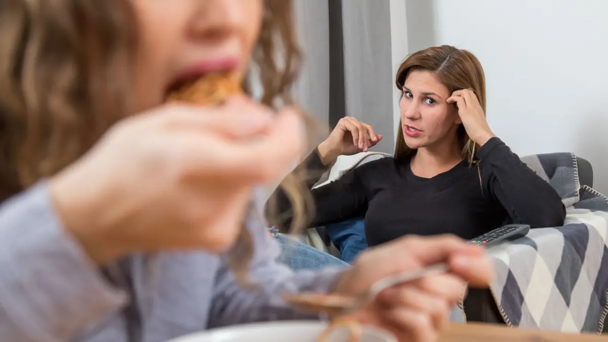 Zwei Frauen im Wohnzimmer, eine Frau isst.: ILLUSTRATION - Menschen mit Misophonie können beispielsweise Kaugeräusche ihrer Mitmenschen kaum ertragen. (zu dpa: «Warum uns bestimmte Geräusche auf die Palme bringen») Foto: Christin Klose/dpa-mag - Honorarfrei nur für Bezieher des Dienstes dpa-Magazin +++ dpa-Magazin +++