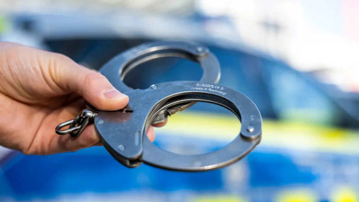 Handschellen vor Polizeiwagen - Symbolbild: ARCHIV - 28.09.2021, Nordrhein-Westfalen, Gütersloh: Eine Hand hält Handschellen vor einen Streifenwagen der Polizei. (zu dpa: «Schüsse auf Bäckerei: Spielt Bandenkonflikt eine Rolle?») Foto: David Inderlied/dpa +++ dpa-Bildfunk +++
