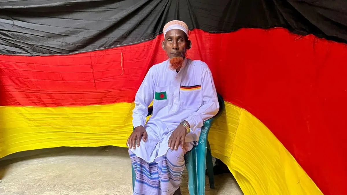 7,5 Kilometer lange deutsche Flagge genäht: PRODUKTION - 14.06.2026, Bangladesch, Magura: Der 77-jährige bangladeschische Bauer Amjad Hossain posiert mit der deutschen Flagge, die er zu Hause genäht hat. Er entrollte die 7,5 Kilometer lange Flagge am 10. Juni auf dem Pausenplatz einer örtlichen Schule, einen Tag vor Beginn der FIFA-Weltmeisterschaft 2026. (zu dpa: «Fan aus Bangladesch näht 7,5-Kilometer-Fahne für DFB-Team») Foto: Nazrul Islam/dpa +++ dpa-Bildfunk +++