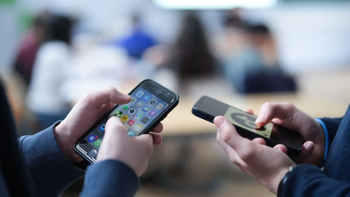 Smartphones in der Schule: ILLUSTRATION - 13.02.2026, Hamburg: Zwei Schüler stehen ein einem Klassenraum und schauen auf ihre Smartphones, auf einem der Displays ist das Logo der Social-Media-Plattform TikTok zu sehen. (zu dpa: «Expertenkommission: Handyverbot in Schulen bis Klasse sieben») Foto: Marcus Brandt/dpa +++ dpa-Bildfunk +++