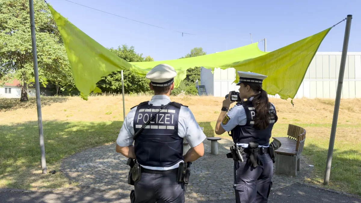 Zwei Polizisten in Uniform stehen unter einem gelben Sonnensegel in einem Park. Sie beobachten die Umgebung, während eine Beamtin ein Gerät in der Hand hält. Im Hintergrund sind Bäume, eine Wiese und ein Gebäude zu sehen.