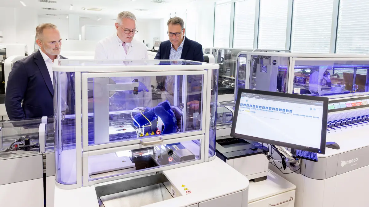 Landrat Richard Sigel (rechts) und Klinik-Geschäftsführer André Mertel (links) lassen sich von Chefarzt Hannes Wickert am Start-Modul der FlexLab-X-Automation zeigen, wie der Roboterarm die Röhrchen greift und auf die Reise schickt.