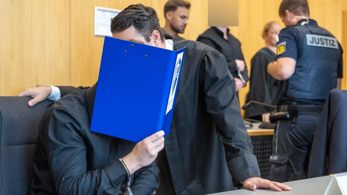Prozess wegen Mordversuch gegen drei junge Männer: 28.08.2026, Baden-Württemberg, Ulm: Im Landgericht bespricht sich ein Mann hinter einer Akte mit seinem Verteidiger, während ein Wachtmeister im Hintergrund einen weiteren Angeklagten bewacht. Die Staatsanwaltschaft wirft drei jungen Männern vor, gemeinsam einen Mordversuch in einem Imbiss begangen zu haben. Foto: Stefan Puchner/dpa +++ dpa-Bildfunk +++