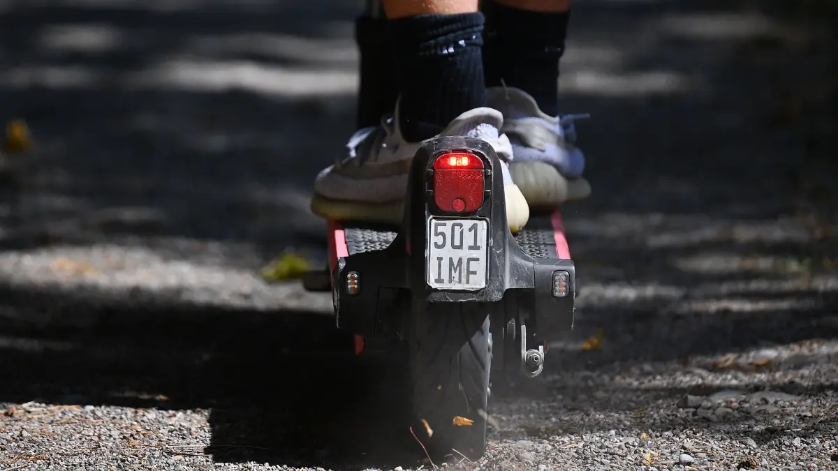 Eine Nahaufnahme vom Heck eines E-Scooters mit Versicherungskennzeichen im Park. Das Bild zeigt eine Detailansicht von hinten auf das Hinterrad eines elektrischen Tretrollers. Am Schutzblech ist ein kleines Versicherungskennzeichen mit der Aufschrift 501 IMF angebracht. Darueber leuchtet ein rotes Ruecklicht. Die Fuesse des Fahrers stehen in hellen Sneakers und schwarzen Socken auf dem Trittbrett. Der Scooter befindet sich auf einem unbefestigten Kiesweg im Schatten. *** A close-up of the rear of an e-scooter with an insurance sticker in the park. The photo shows a detailed view from behind of the rear wheel of an electric scooter. A small insurance sticker with the inscription 501 IMF is attached to the fender. A red taillight shines ab