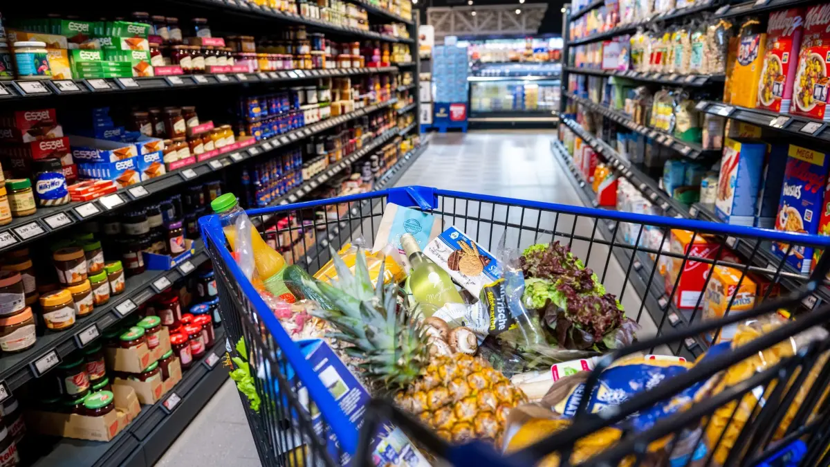 Einkaufswagen mit verschiedenen Lebensmittel zwischen Regalen: ILLUSTRATION - Häufig befinden sich im Supermarkt die günstigeren Produkte weit unten oder weit oben im Regal. Teurere Ware ist hingegen meist auf Augenhöhe. (zu dpa: «Große Packung, kleiner Preis? Drei Spartipps für den Einkauf») Foto: Benjamin Nolte/dpa-mag - Honorarfrei nur für Bezieher des Dienstes dpa-Magazin +++ dpa-Magazin +++