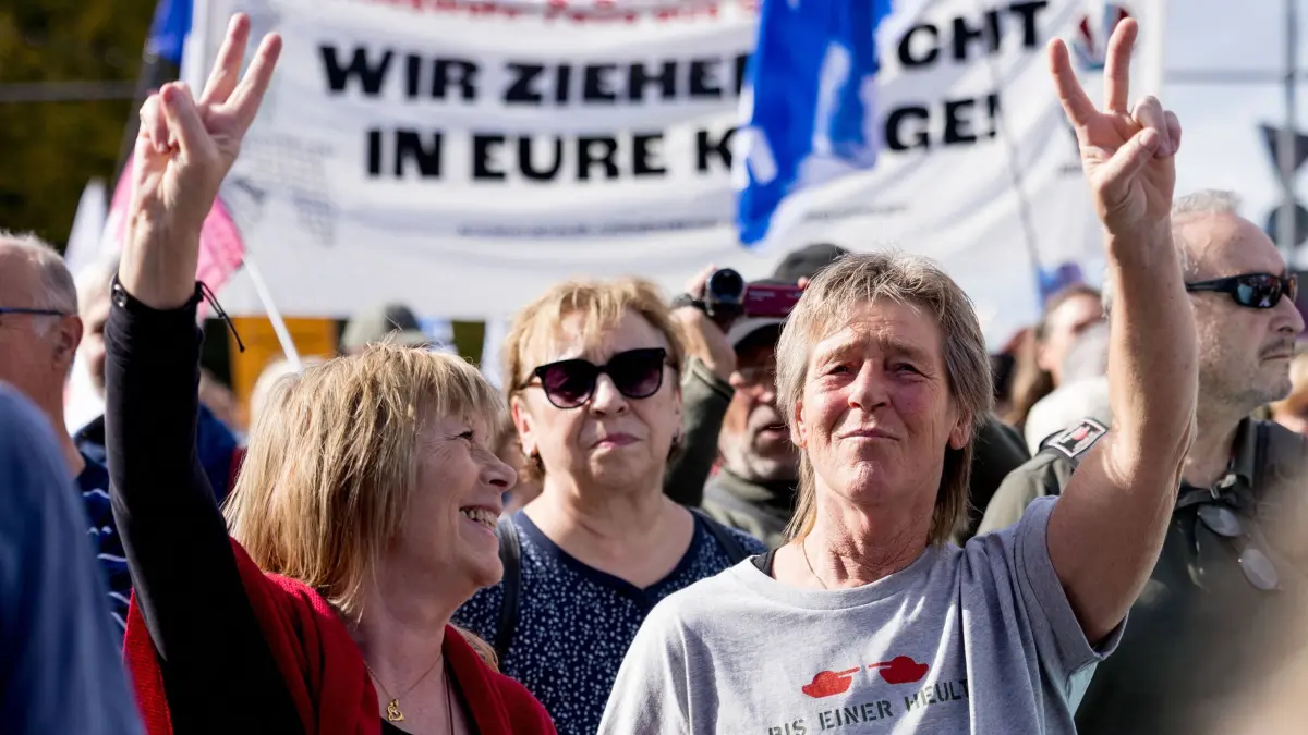 Friedensdemonstration gegen Aufrüstung und Krieg in Berlin: 03.10.2026, Berlin: Ein Banner mit der Aufschrift «Wir ziehen nicht in eure Kriege!» ist bei der Friedensdemonstration «Nein zu Hochrüstung, Wehrpflicht und Krieg!» vor dem Brandenburger Tor zu sehen, während Teilnehmende das Victory-Zeichen zeigen. In mehreren deutschen Städten kommen Menschen zusammen, um gegen Aufrüstung und Krieg zu demonstrieren. Foto: Carsten Koall/dpa +++ dpa-Bildfunk +++