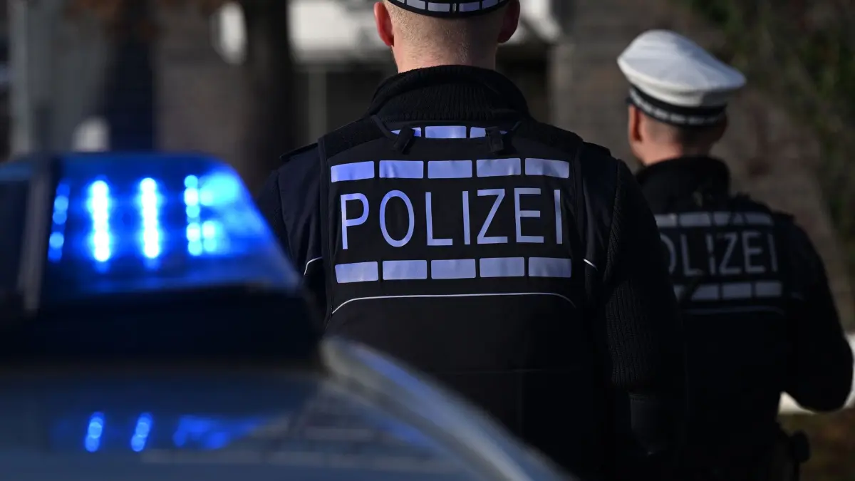 Polizei: Das Logo der Polizei auf der Dienstkleidung von Polizisten die vor einem Blaulicht stehen (gestellte Szene). (zu dpa: «3,5 Kilogramm Kokain entdeckt - Drei Männer in U-Haft»)