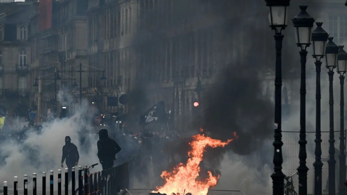 Proteste in Bordeaux: 24.10.2026, Frankreich, Bordeaux: Ein Demonstrant geht an einem Feuer in der Nähe der Place de la Bourse vorbei, die in einer Demonstrationssperrzone liegt, während einer Demonstration für Kaufkraft, den Schutz öffentlicher Dienstleistungen sowie steuerliche und soziale Gerechtigkeit im Rahmen eines landesweiten Aufrufs zur Mobilisierung. Foto: Philippe Lopez/AFP/dpa +++ dpa-Bildfunk +++
